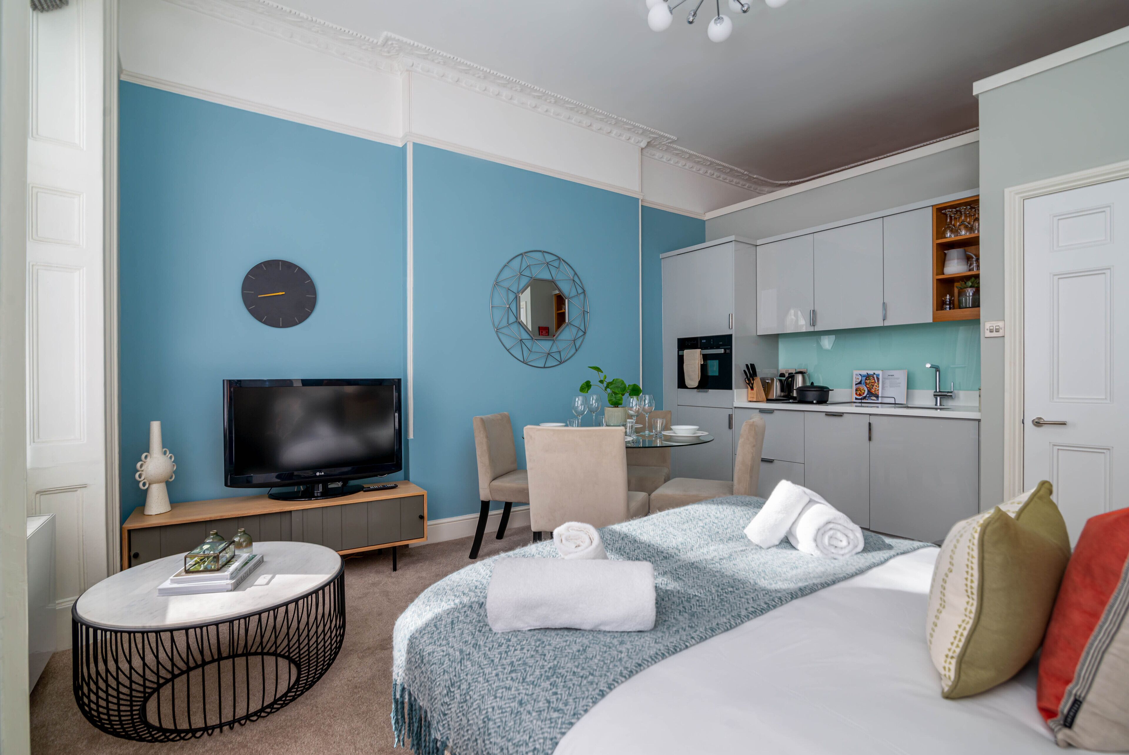 A modern hotel room with a TV, a round coffee table, a bed with a blue patterned bedspread, a dining area with a table and chairs, and a kitchen area with cabinets and a countertop.