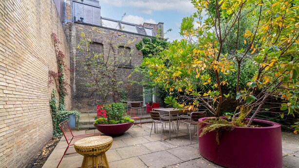 Une cour avec un mur en briques, un grand bac à fleurs contenant un arbre, du mobilier d’extérieur et une chaise rouge.