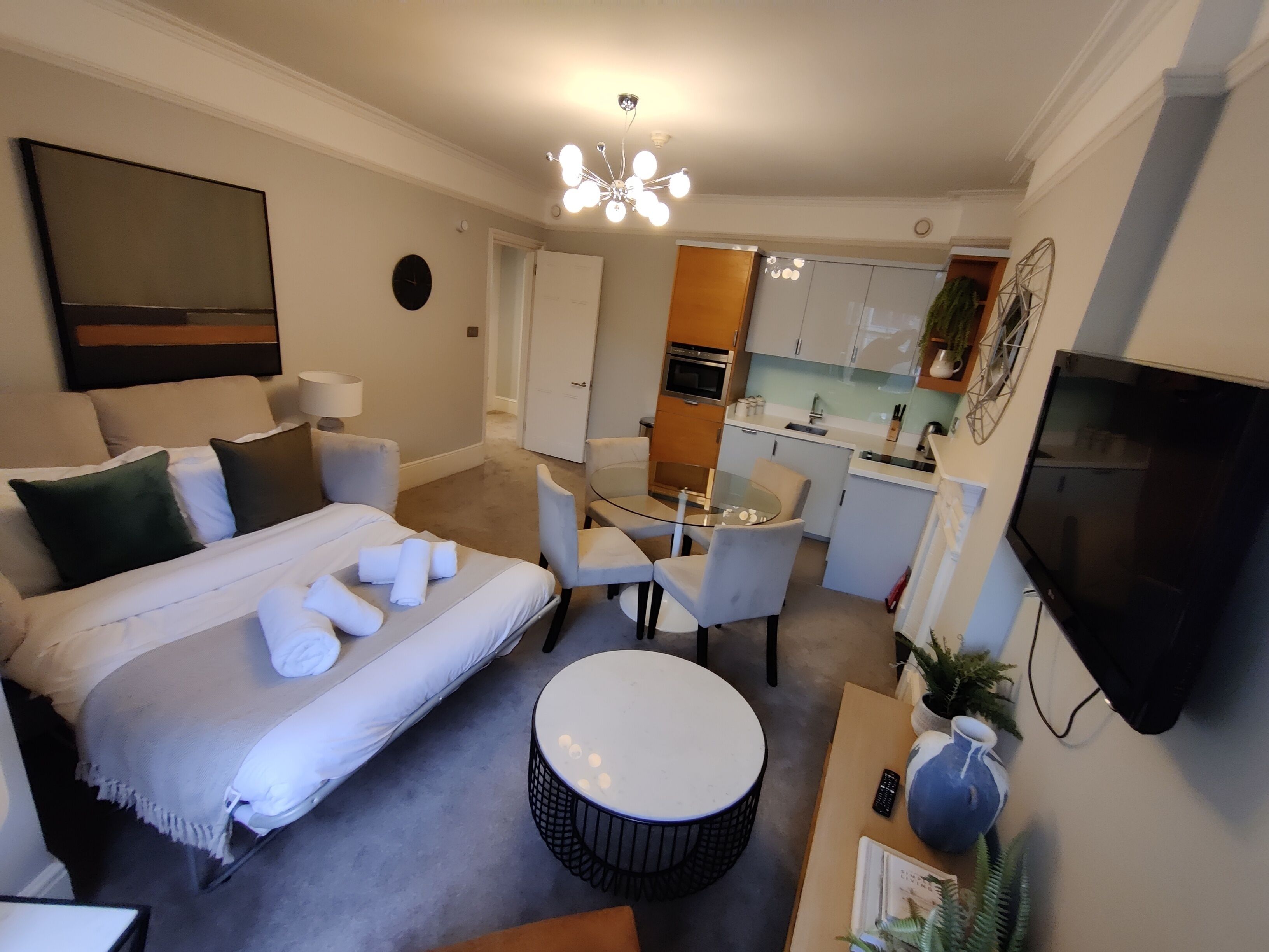 A modern hotel room with a bed, seating area, kitchenette, and a TV.