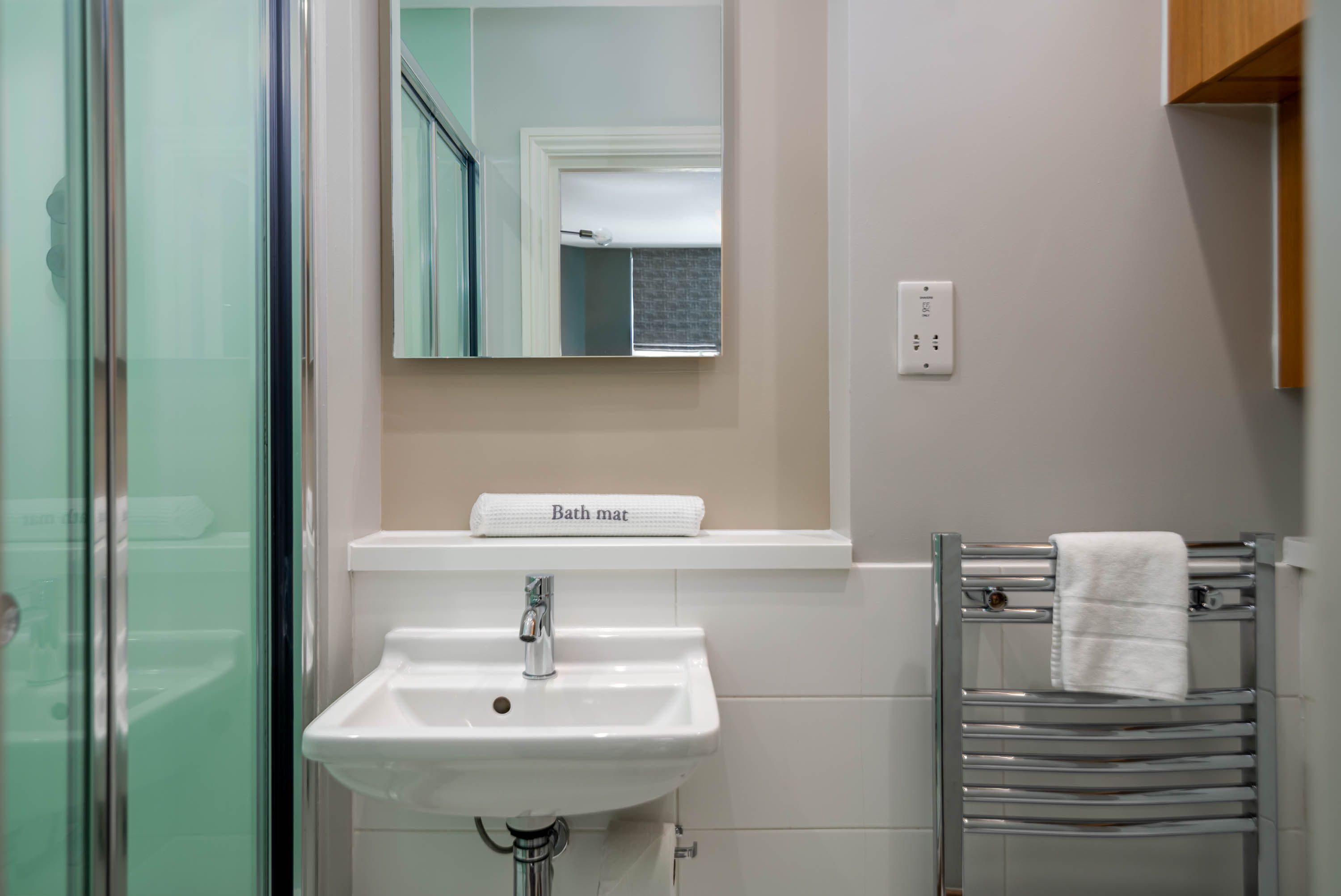 Un baño con cabina de ducha de vidrio, un lavamanos blanco, un espejo y un perchero para toallas.