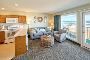 A compact living space with a kitchenette, a gray sofa, a round wicker ottoman, and a view of the ocean through a sliding glass door.