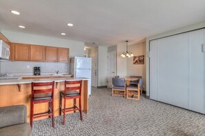 A compact kitchen with wooden cabinets, a refrigerator, a coffee maker, and a dining area with two chairs and a table. - Beachfront Family Suite - Ocean View - Full Kitchen (Lincoln City)