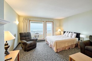 A hotel room with a bed, two armchairs, a desk with a lamp, a view of the sea through a window, and a balcony.