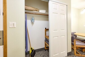 Een kamer met een stapelbed, een klein tafeltje met stoelen, een bezem en een wandradiator.
