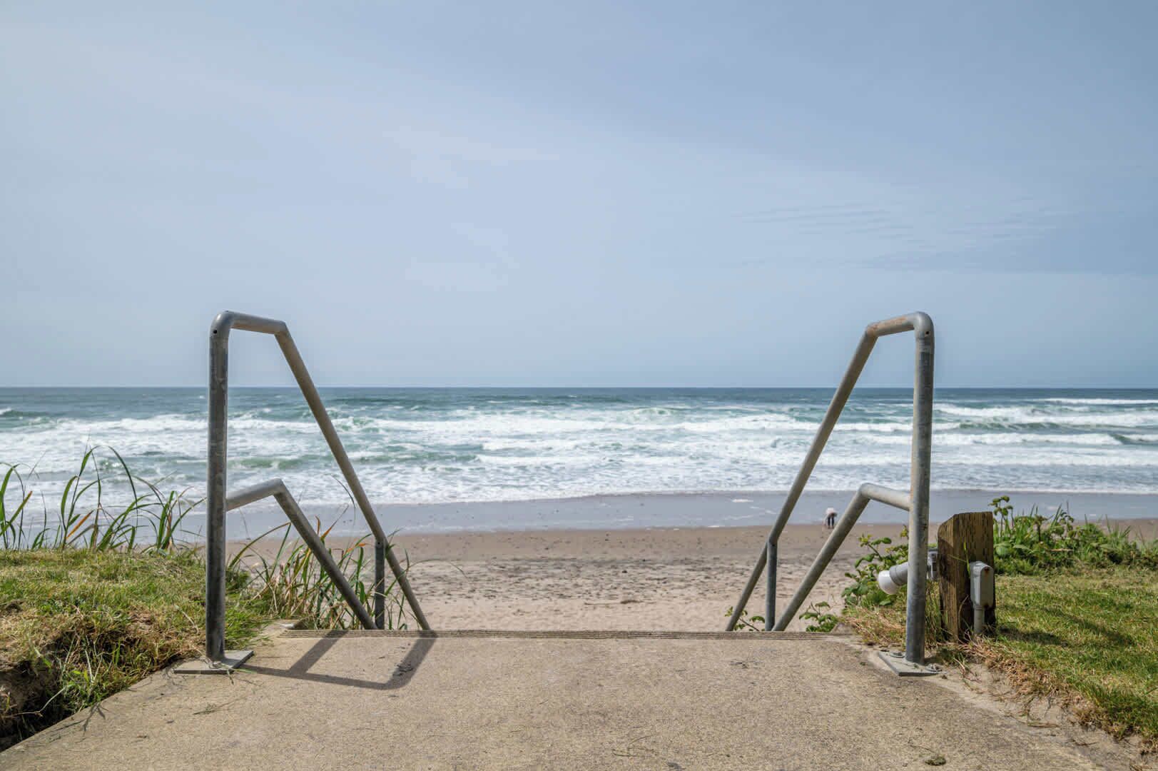 A concrete pathway leading to a beach with metal railings and ocean waves in the background.