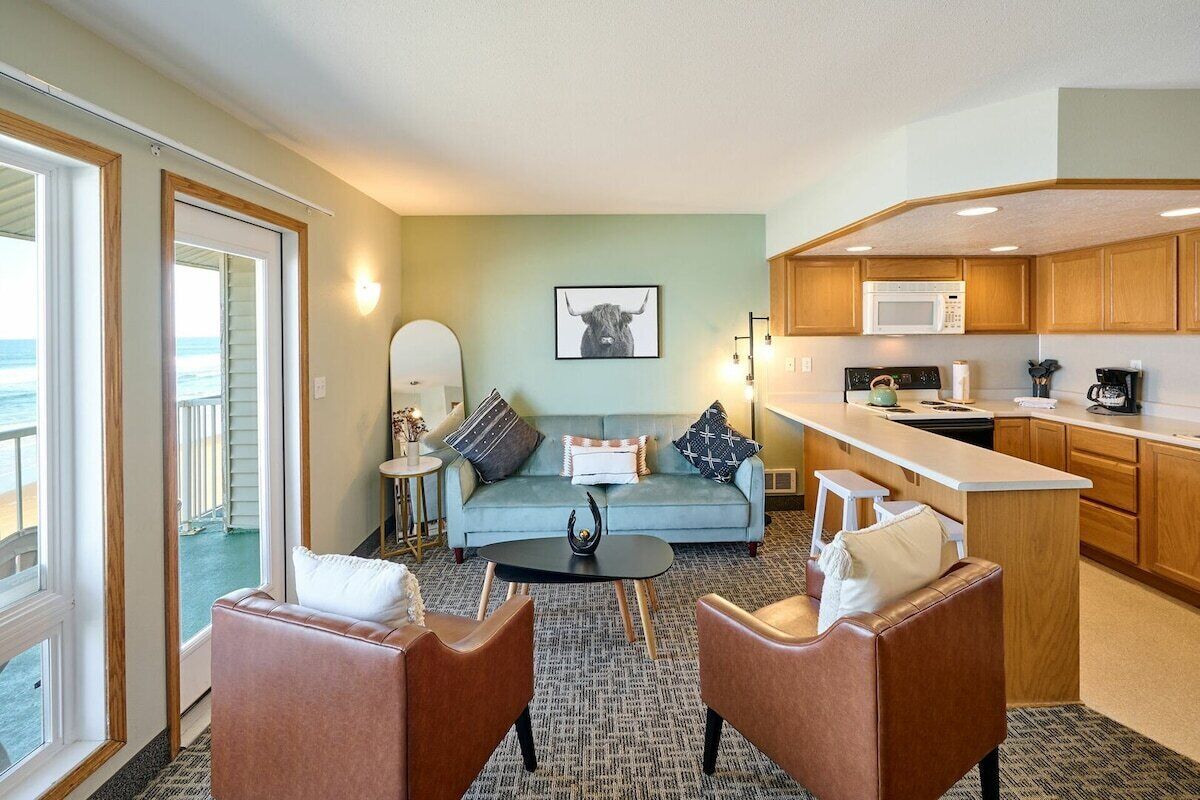 A compact living space with a sofa, two armchairs, a coffee table, a kitchen area with a microwave, and a view of the ocean through a sliding door.