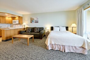 Een hotelkamer met kitchenette, een bed met witte dekbedovertrek, een bank met gestreepte kussens, een eettafeltje en twee bedlampjes.