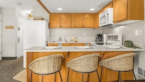 A kitchen with wooden cabinets, a white refrigerator, a microwave, an oven, a sink, and three woven bar stools. - Oceanfront Family Suite - Full Kitchen - Beautiful Views and Pool & Hot Tub Access! (Lincoln City)