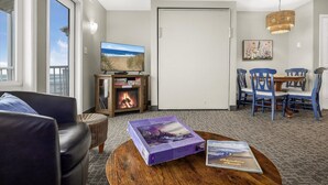 A living room with a fireplace, a television, a dining area with blue chairs, and a view of the ocean through a large window. - Oceanfront Family Suite - Full Kitchen - Beautiful Views and Pool & Hot Tub Access! (Lincoln City)