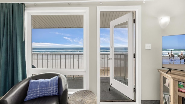 A room with a view of the beach, a balcony, a leather chair, a wicker ottoman, and a television. - Oceanfront Family Suite - Full Kitchen - Beautiful Views and Pool & Hot Tub Access! (Lincoln City)
