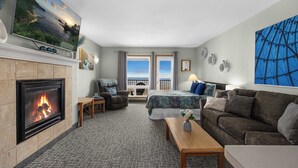 A hotel room with a fireplace, a bed with a view, a sofa, a table, and a large painting. - Top Floor Ocean View Suite - Sleeps Six - Pool & Hot Tub Access! (Lincoln City)