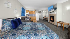 A hotel room with a large bed, a fireplace, a dining area, and a kitchen in the background. - Top Floor Ocean View Suite - Sleeps Six - Pool & Hot Tub Access! (Lincoln City)