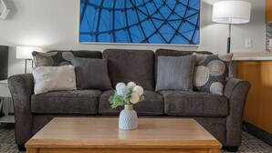 A modern living room with a brown sofa, a white vase with flowers, a wooden coffee table, and a large abstract painting on the wall. - Top Floor Ocean View Suite - Sleeps Six - Pool & Hot Tub Access! (Lincoln City)