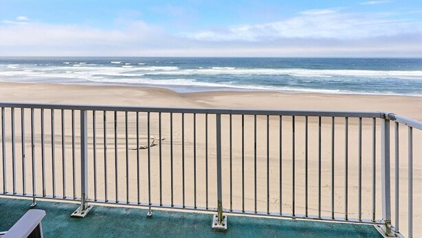 Vista a la playa desde un balcón con barandilla de metal, cielo despejado y el océano al fondo.