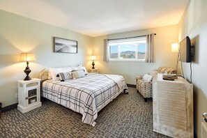A hotel room with a bed, bedside tables, a TV, a window with curtains, and a view of the outside. - Amazing Ocean View Suite - Sleeps Six - Pool Access & Hot Tub! (Lincoln City)