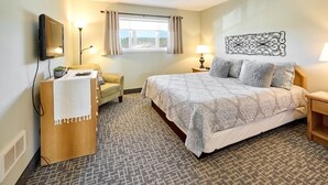 A hotel room with a bed, a desk with a lamp, a chair, a TV, and a window with curtains. - Ocean View Suite - Sleeps Six - Heated Indoor Pool & Hot Tub Access (Lincoln City)