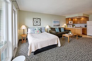 A compact hotel room with a bed, a sofa, a small kitchen area, and a view through a window.