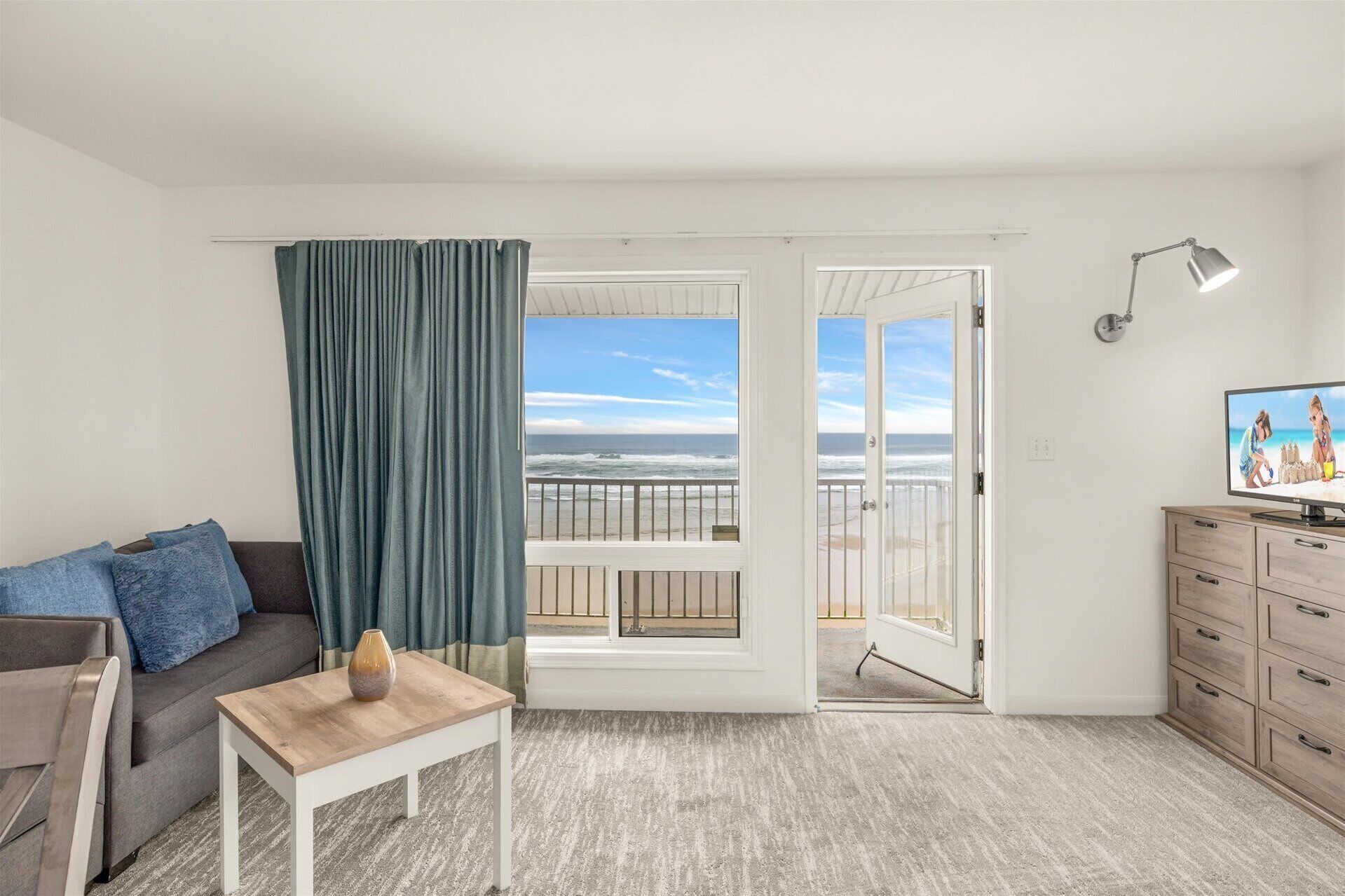 Et værelse med sofa, et lille bord, et fjernsyn, en balkon med udsigt til stranden og en lampe.