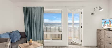 Et værelse med sofa, et lille bord, et fjernsyn, en balkon med udsigt til stranden og en lampe.