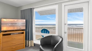 Une chambre dotée d’une télévision, d’une commode en bois, d’une chaise noire et d’un balcon offrant une vue sur la plage.