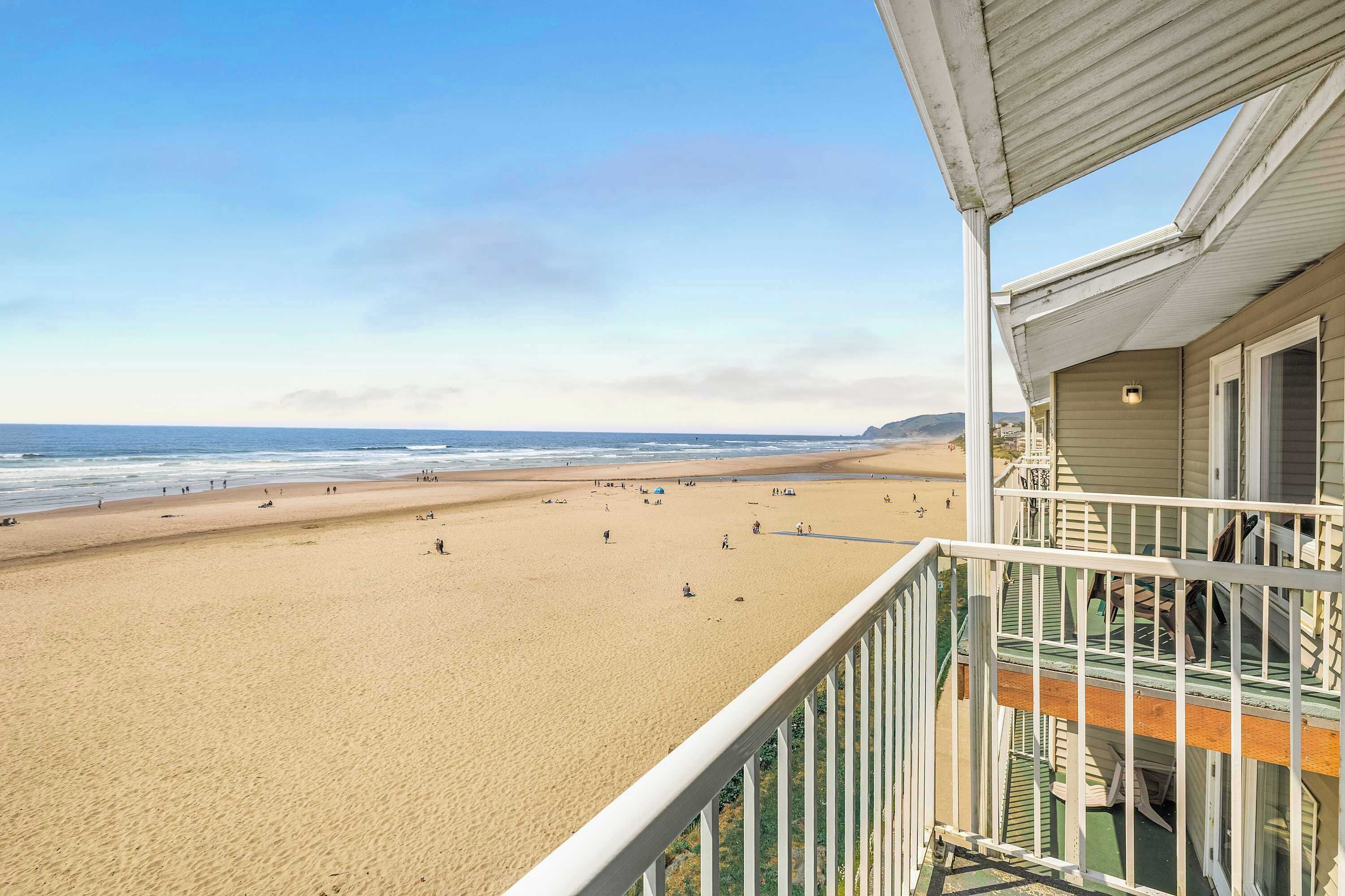 Une vue sur la plage depuis un balcon doté d’une balustrade, un ciel dégagé et une plage de sable où se trouvent des personnes.