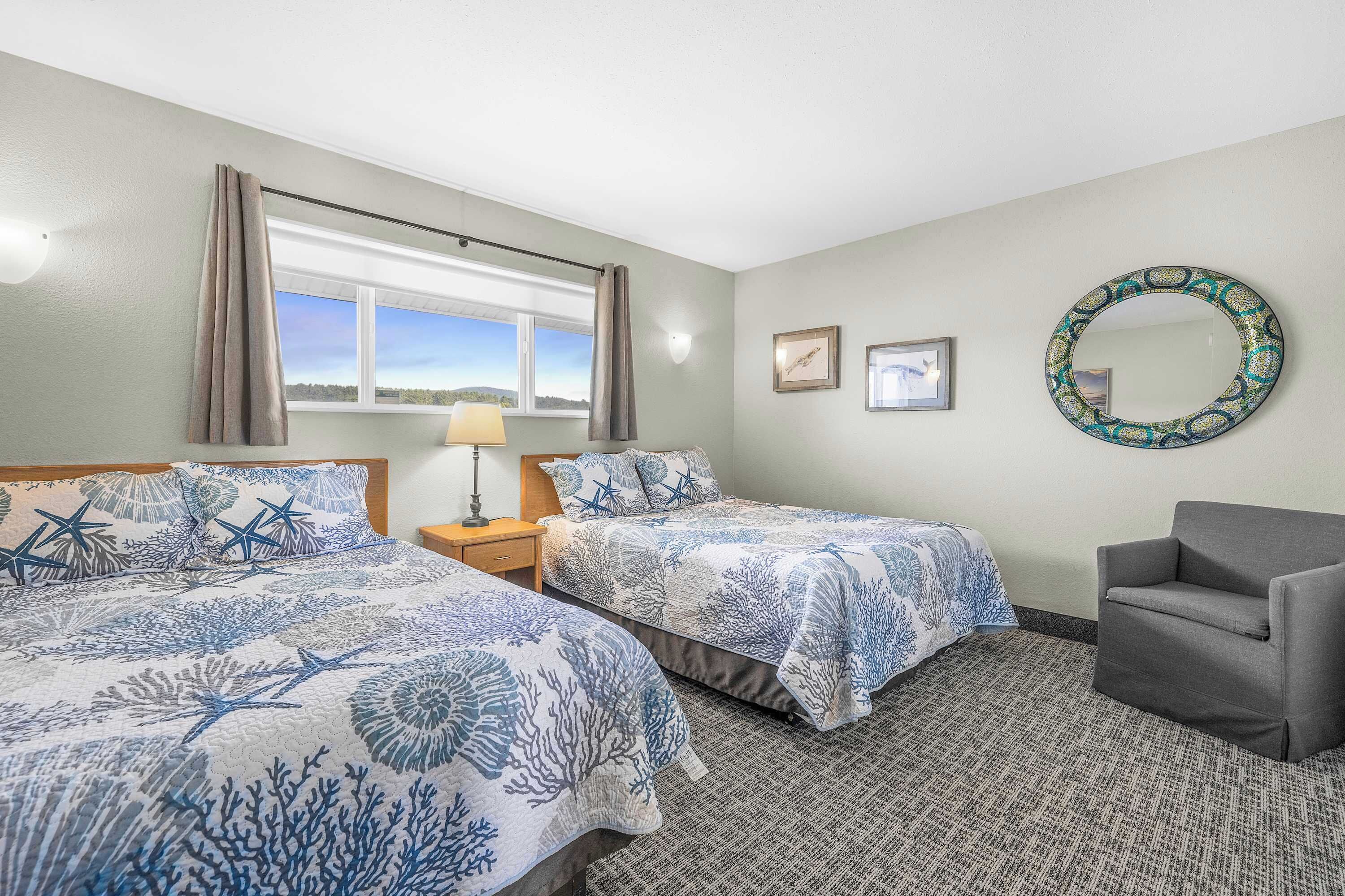 A hotel room with two beds, a gray armchair, a window with curtains, a wall-mounted mirror, and framed pictures.