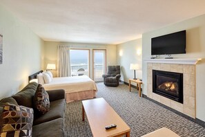 A hotel room with a bed, a sofa, a fireplace, a television, and a balcony with curtains. - Fireplace Suite With King Bed and Full Kitchen - Pool and Hot Tub Access! (Lincoln City)