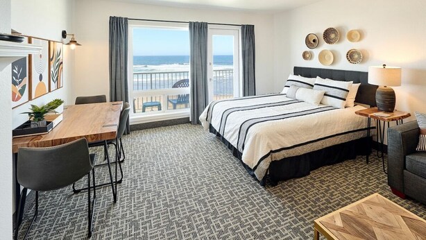 A hotel room with a bed, a desk, a chair, a small table, and a view of the ocean through a window.