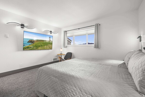 Un dormitorio con una cama grande, un escritorio, una silla, un ventanal con cortinas y un cuadro de un paisaje costero.