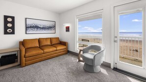A modern living room with a brown leather sofa, a gray armchair, a small table, and a large window with a beach view. - Corner King Suite, Stunning Ocean and River Views! (Lincoln City)