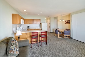 A compact living space with a kitchenette, a dining area with chairs, a sofa with a patterned cushion, and a carpeted floor.
