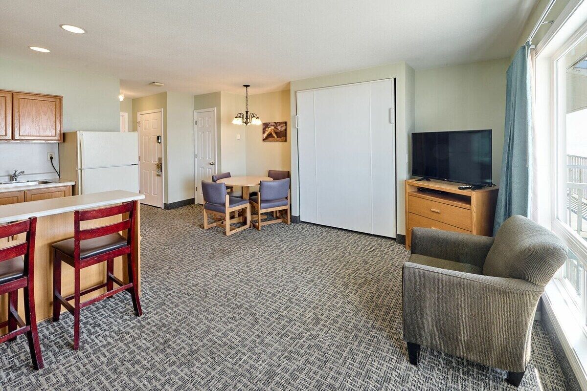 Habitación de hotel compacta con kitchenette, comedor con mesa y sillas, televisión y ventana grande.
