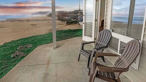 Uitzicht op het strand vanaf een veranda met twee Adirondack-stoelen, een betonnen terras en een glazen deur die naar een kamer leidt.