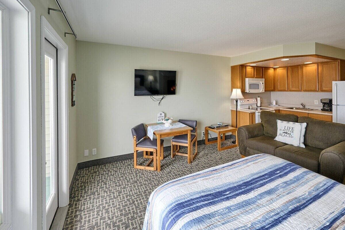 Quarto de hotel com cama, sofá, pequena mesa de jantar, televisão e cozinha compacta.
