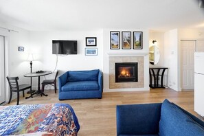 A room with a fireplace, a television mounted on the wall, a blue sofa, a dining table with chairs, and a bed with a colorful blanket.