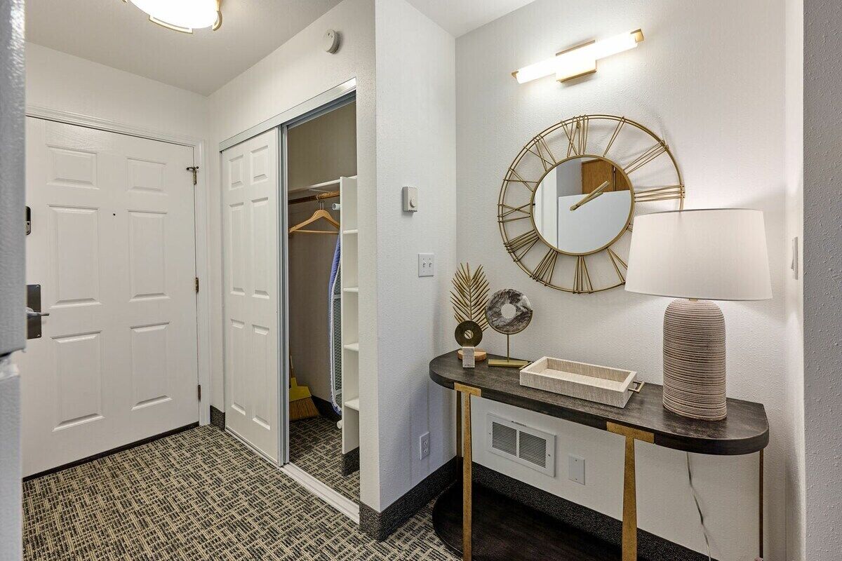 A hallway with a coat closet, a round mirror, and a table with a lamp and decorative items.