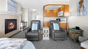 A cozy living room with a fireplace, two leather armchairs, a coffee table, and a kitchen area with wooden cabinets and a microwave. - Ground Floor Studio - Ocean View - Pool & Hot Tub! (Lincoln City)