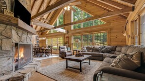 A log cabin interior with a stone fireplace, leather furniture, and large windows.