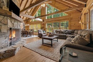 A log cabin interior with a stone fireplace, leather furniture, and large windows. - Hot Tub, Game Room & Valley Views + Biltmore Pass | Peaceful Mountain Lodge (Burnsville)