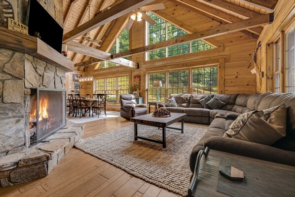 A log cabin interior with a stone fireplace, leather furniture, and large windows.