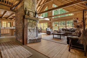 A spacious living area with a stone fireplace, wooden beams, and large windows. - Hot Tub, Game Room & Valley Views + Biltmore Pass | Peaceful Mountain Lodge (Burnsville)