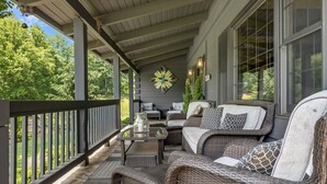 A covered porch with wicker furniture, a decorative wall piece, and a view of greenery.