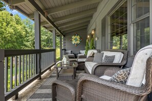 A covered porch with wicker furniture, a decorative wall piece, and a view of greenery.