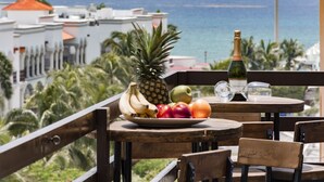 Una mesa con una bandeja de fruta, una botella de vino y vasos en un balcón con vista al océano.