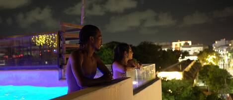 Vista nocturna de un área de piscina con barandilla, palmeras y luces de la ciudad a lo lejos.