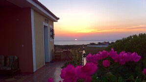Una terraza con vista a la puesta de sol sobre el mar, una silla y flores rosadas en primer plano.
