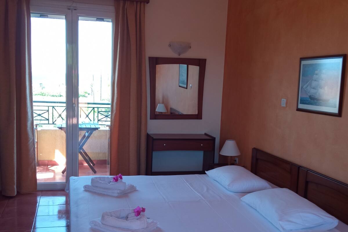 A hotel room with a large bed, a wooden headboard, a nightstand, a lamp, a framed picture on the wall, and a sliding glass door leading to an outdoor area.