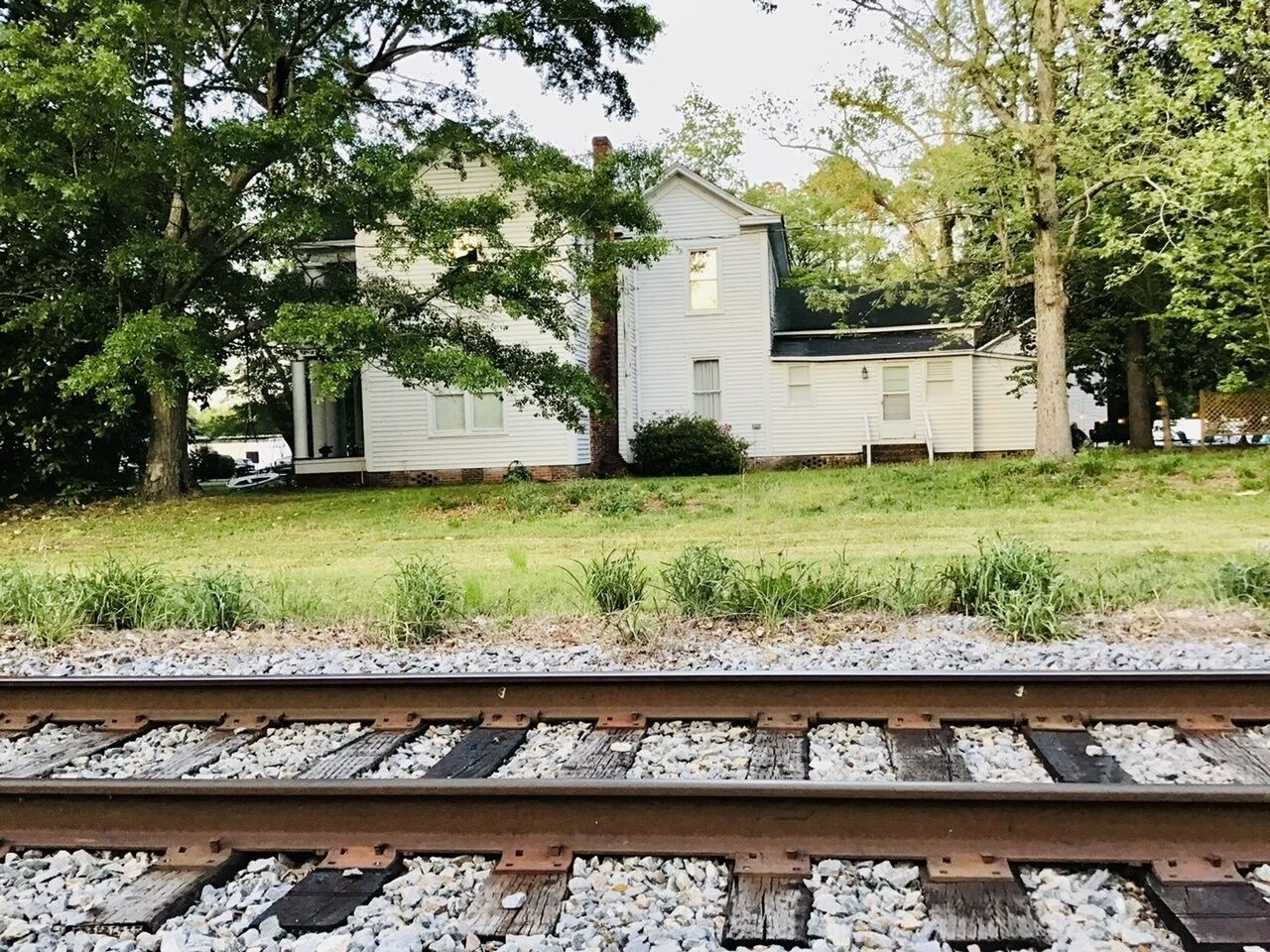 A house with a small porch and a garage, surrounded by trees and grass, with a railway track in the foreground.