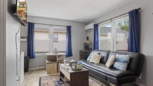 Een moderne woonkamer met een leren bank, een salontafel en een klein bureau. Twee ramen met blauwe gordijnen, een airconditioning en uitzicht op het buiten.