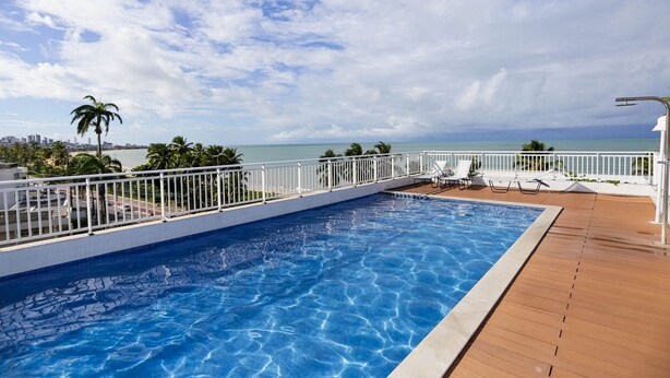 A swimming pool with a wooden deck and white railing, overlooking a beach and ocean. - Apart Praia de Cabo Branco por Carpediem (João Pessoa)